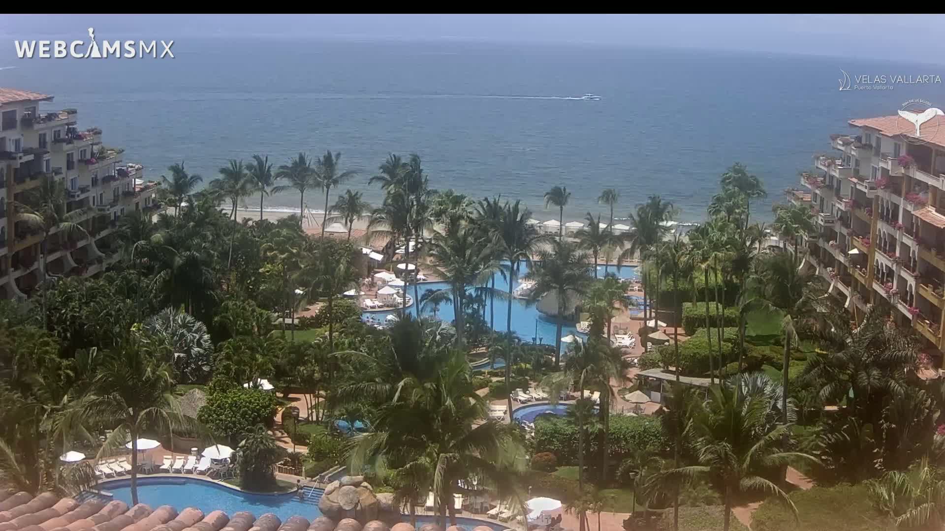 A tropical resort with multiple pools is situated between lush greenery and a calm ocean under a sunny sky.