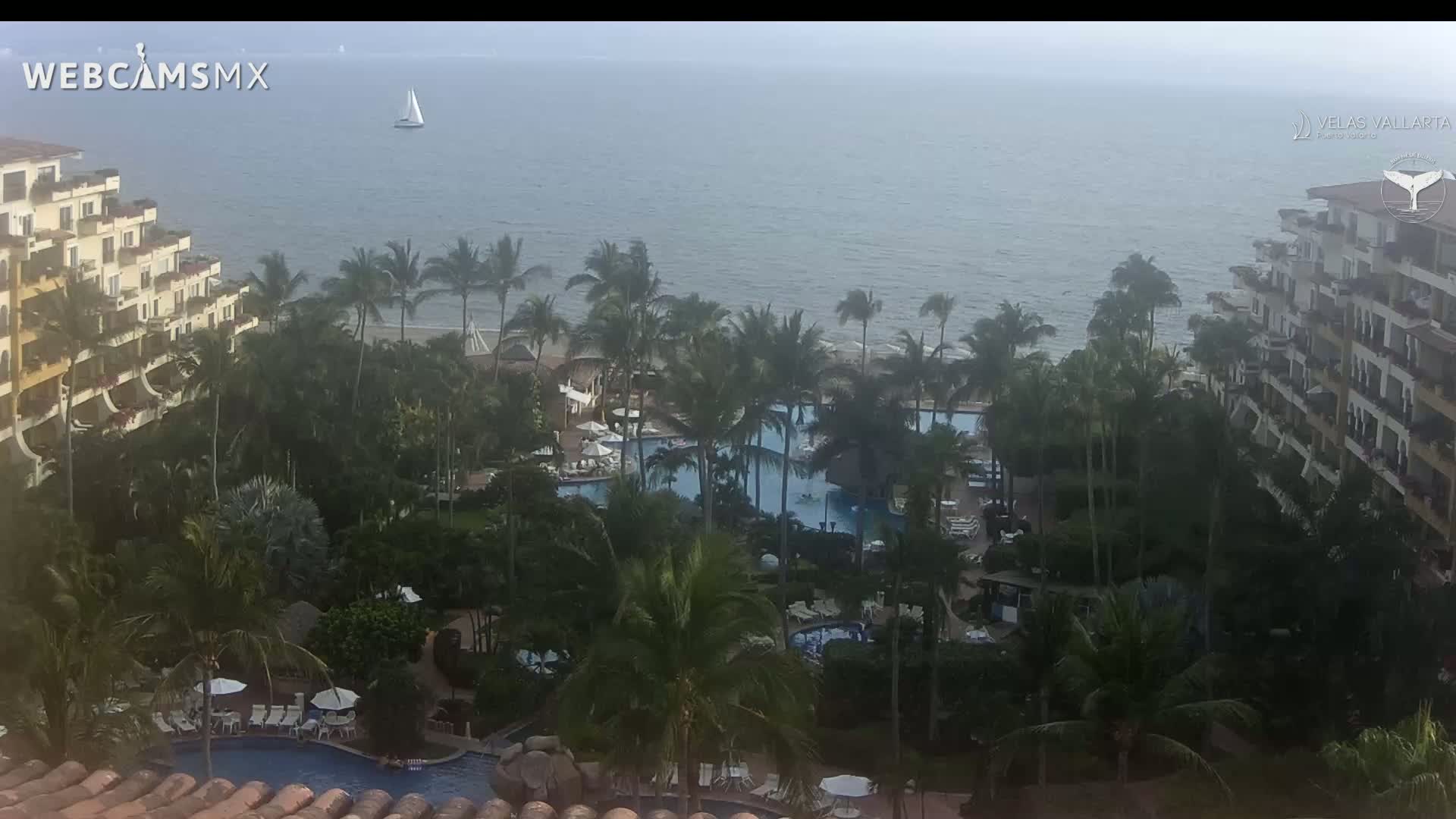 An elevated view shows a resort with multiple pools surrounded by lush tropical vegetation, overlooking a calm ocean with a sailboat in the distance under an overcast sky.