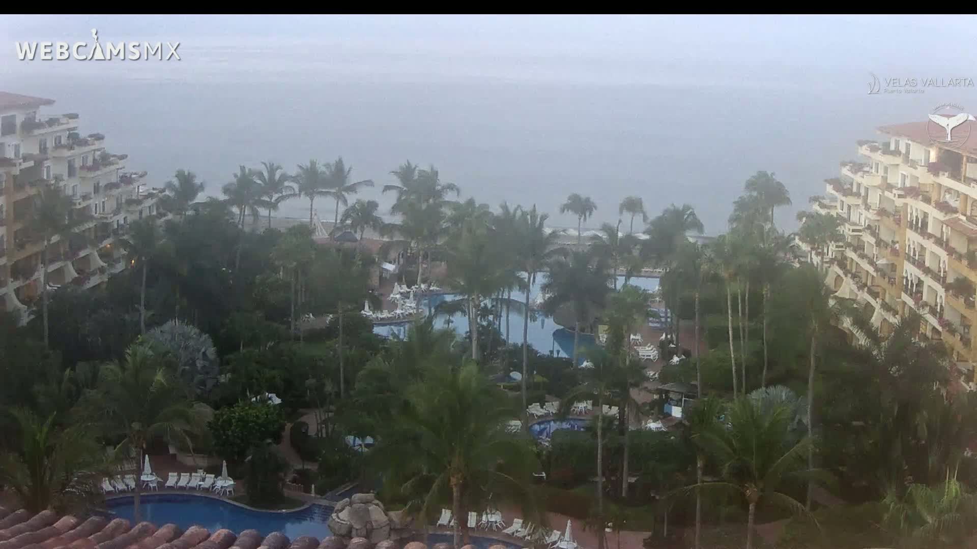A hazy view shows a tropical resort with multiple pools surrounded by palm trees and large buildings, overlooking a foggy ocean.