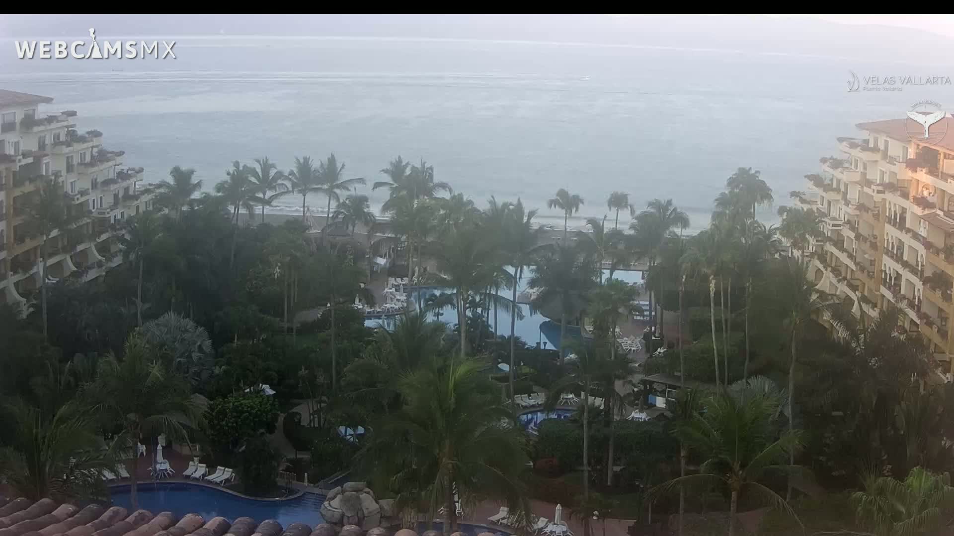 A hazy, overcast day reveals a resort with multiple pools surrounded by lush tropical vegetation and buildings, overlooking a calm ocean.