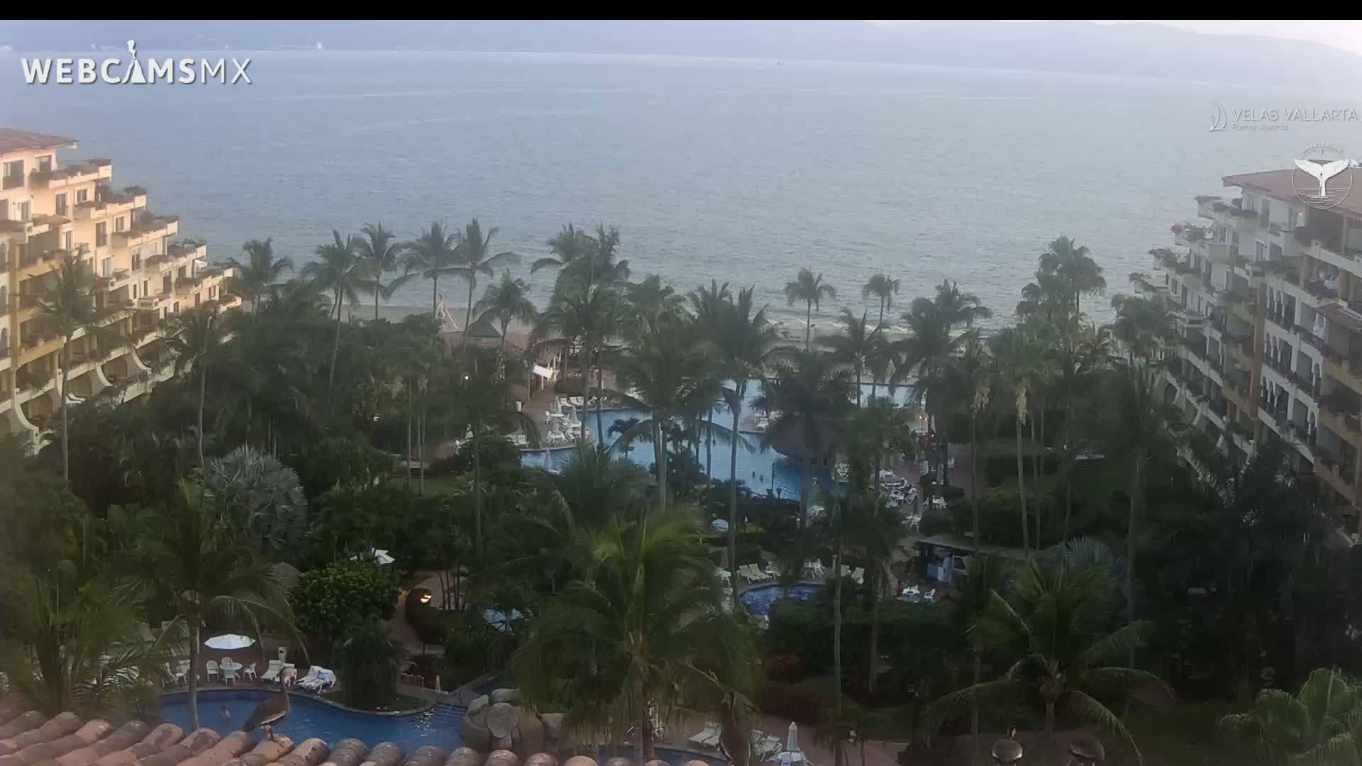 An aerial view shows a tropical resort with multiple pools, palm trees, and beachfront buildings overlooking a calm ocean under a cloudy sky.