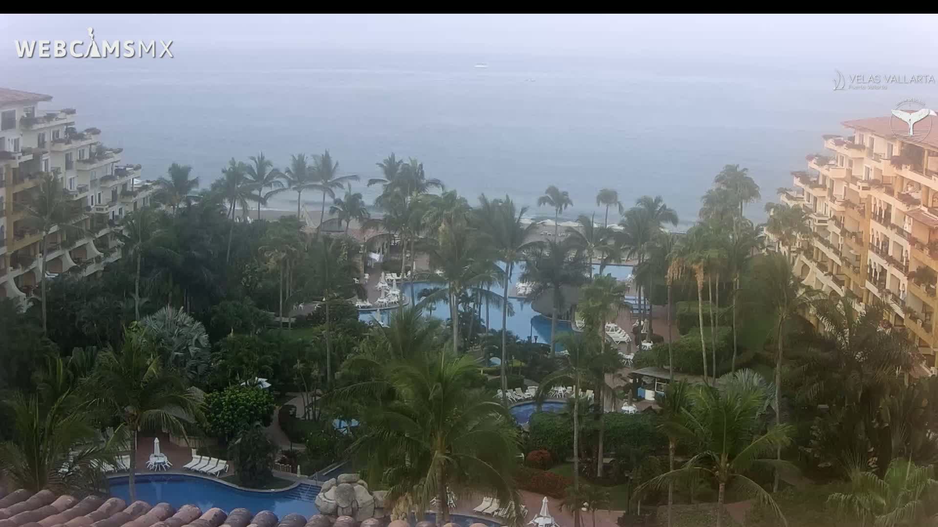 An aerial view shows a resort with multiple pools surrounded by lush tropical vegetation and overlooking a hazy ocean.