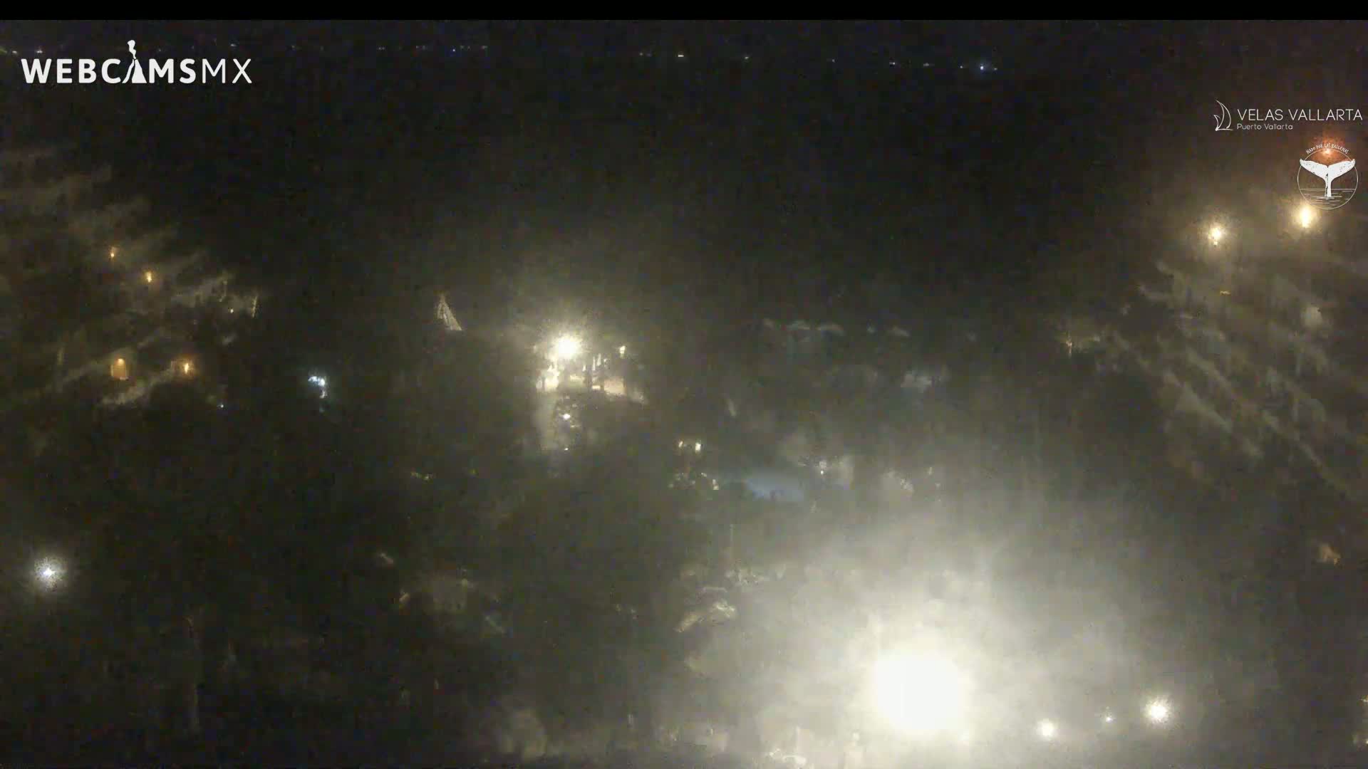 A nighttime aerial view shows a dimly lit area with scattered lights, possibly buildings or structures, under hazy or foggy conditions.