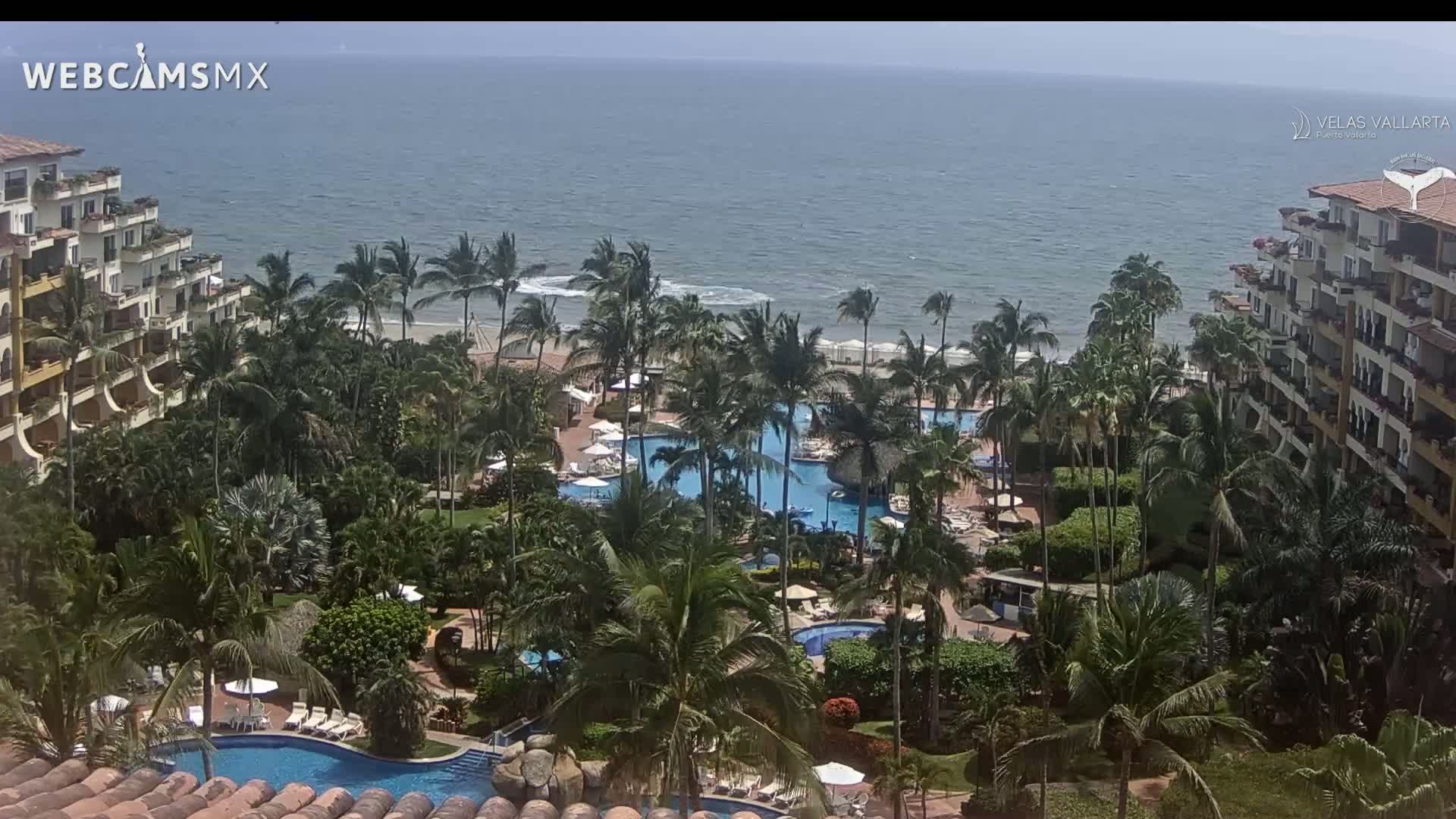 A resort with multiple pools surrounded by palm trees overlooks a beach and ocean on a partly sunny day with light winds.