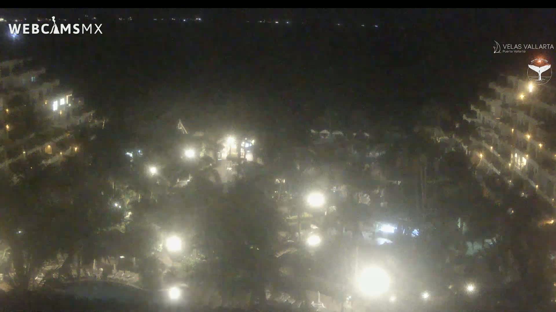 A nighttime, high-angle view shows a resort complex with numerous buildings lit by outdoor lights, surrounded by dark vegetation.