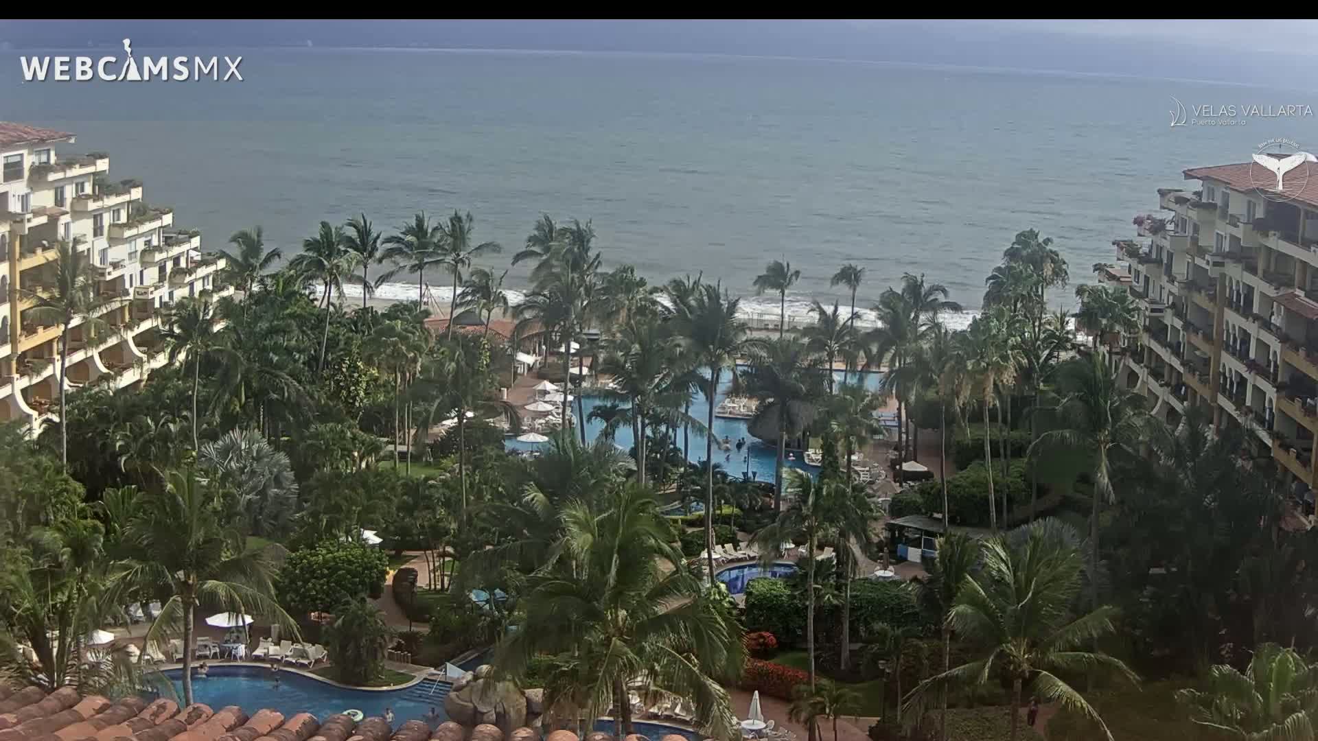 An aerial view shows a tropical resort with multiple pools surrounded by palm trees and buildings overlooking a somewhat cloudy ocean.