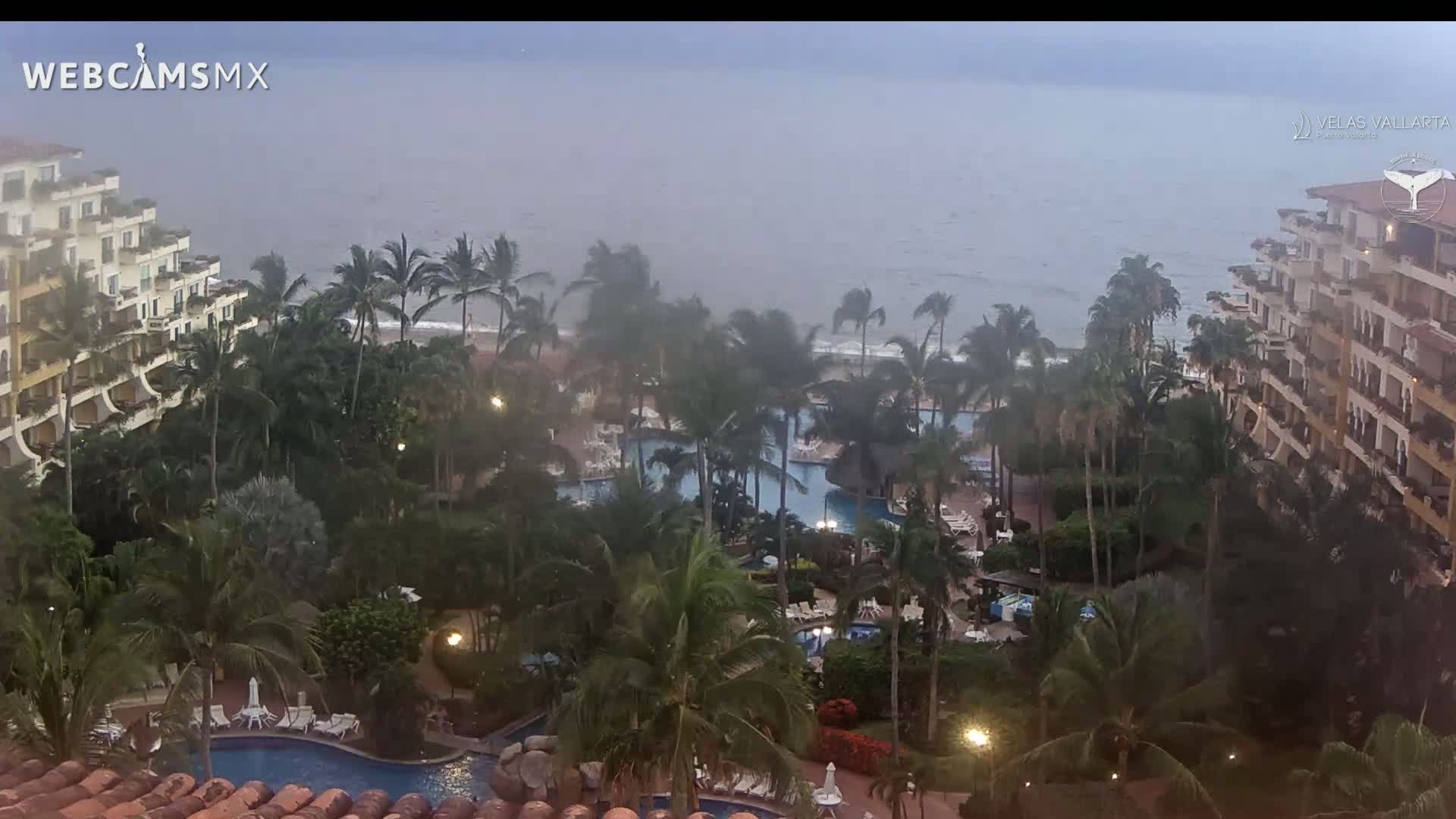 An aerial view shows a resort with multiple pools surrounded by palm trees and buildings overlooking a stormy ocean.