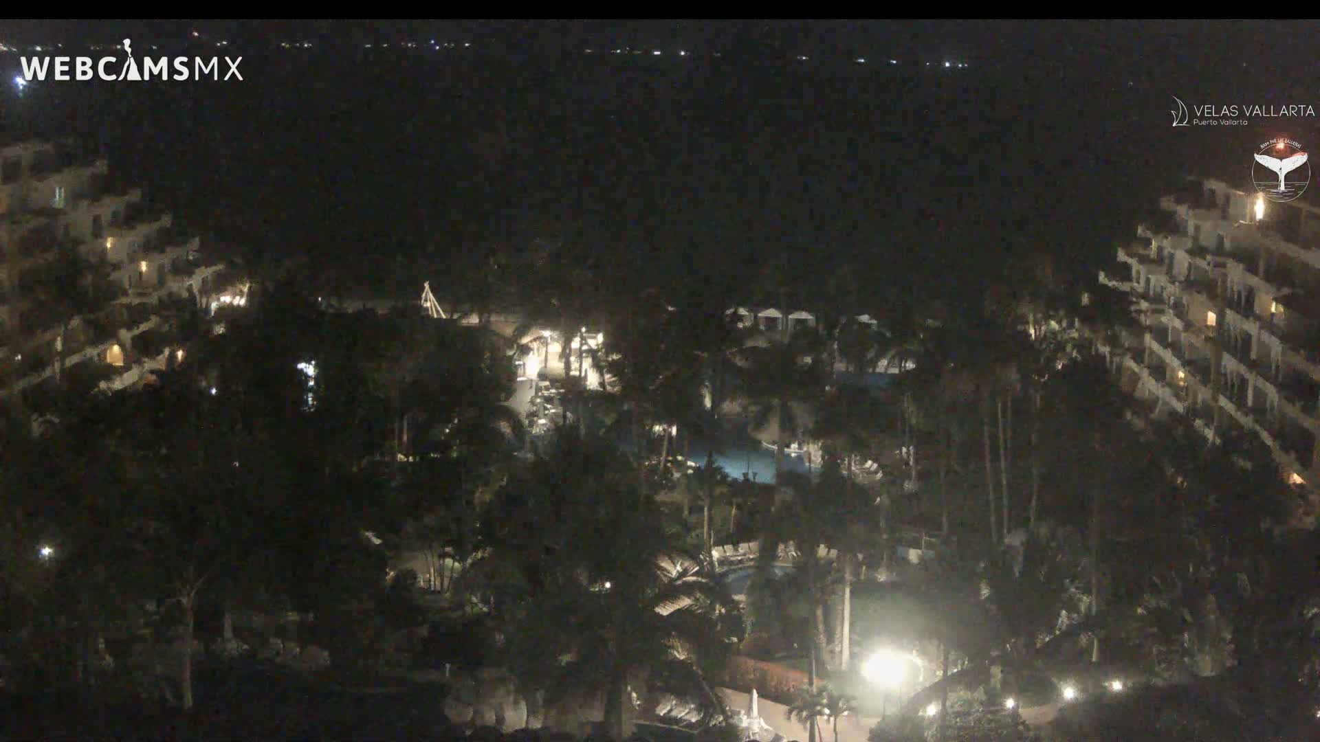 A nighttime aerial view shows a resort complex with pools and palm trees, illuminated by artificial lighting.