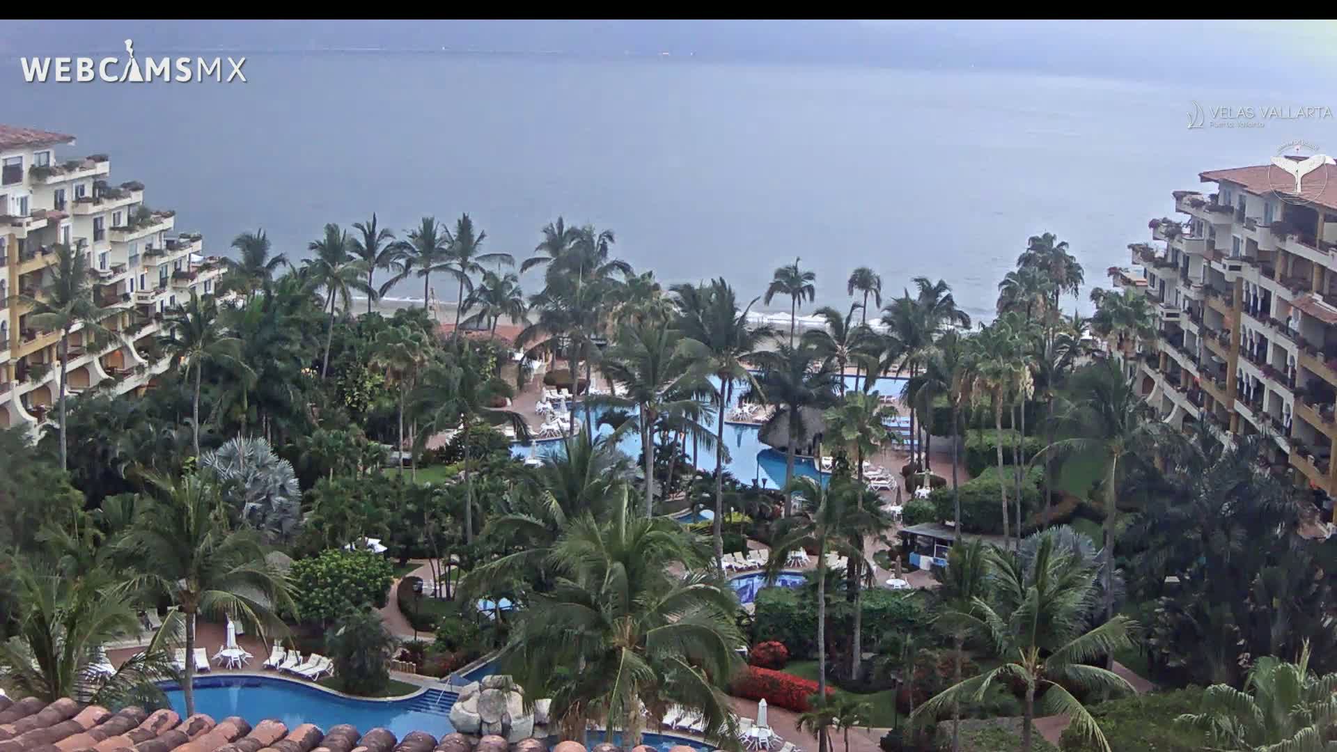 An overcast day reveals a resort with multiple pools surrounded by lush tropical landscaping and palm trees, overlooking a calm ocean.