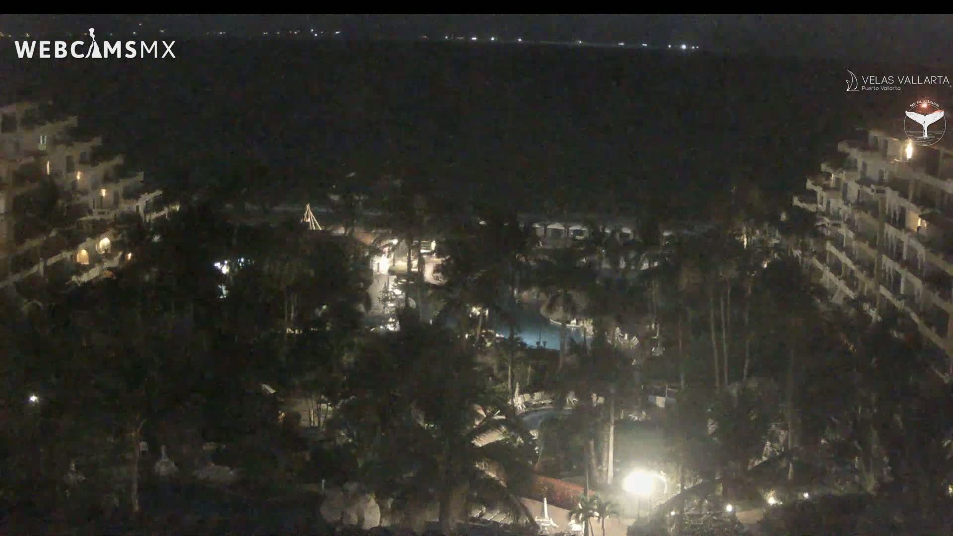 A nighttime, high-angle view of a resort complex with palm trees, pools, and buildings illuminated by lights, overlooking a dark ocean.