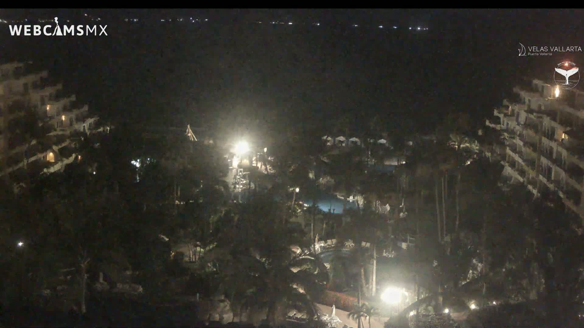 A nighttime aerial view shows a resort complex with pools and palm trees, illuminated by artificial lights, overlooking a dark body of water under a clear sky.