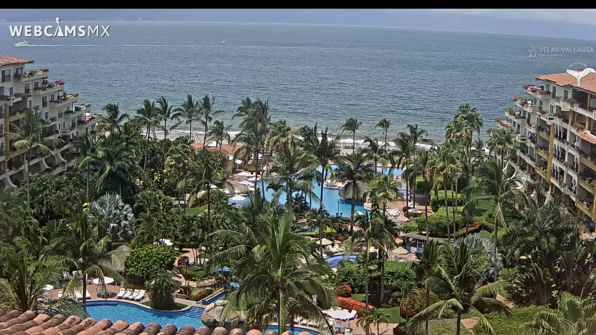 An aerial view shows a tropical resort with multiple pools surrounded by palm trees and buildings, overlooking a calm ocean under a partly cloudy sky.