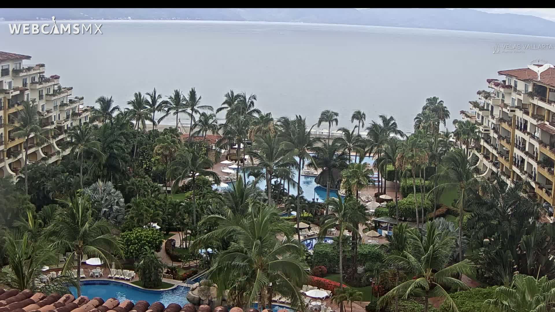 An overcast day reveals a tropical resort with multiple pools surrounded by lush palm trees and multi-story buildings overlooking a calm ocean.