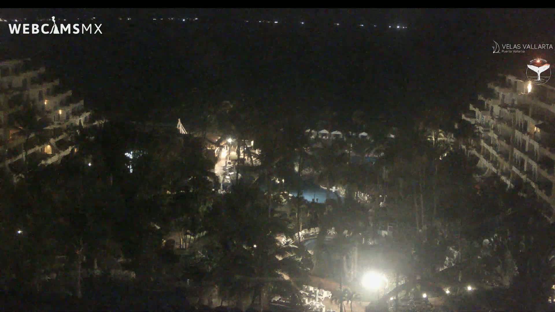 A nighttime aerial view shows a resort complex with pools and buildings surrounded by lush, dark foliage, overlooking a calm, dark body of water under a clear sky.