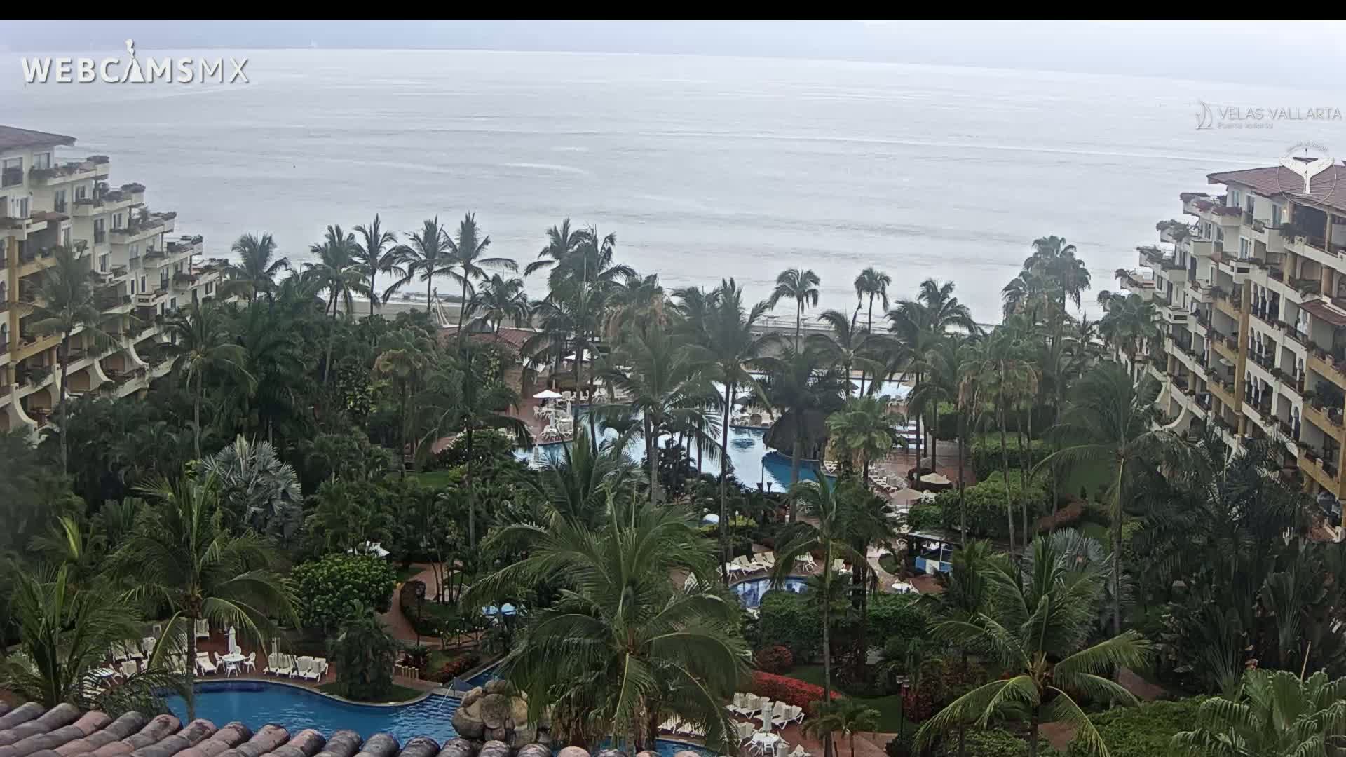 An overcast day reveals a resort with multiple pools surrounded by lush tropical vegetation and beachfront buildings overlooking a calm ocean.
