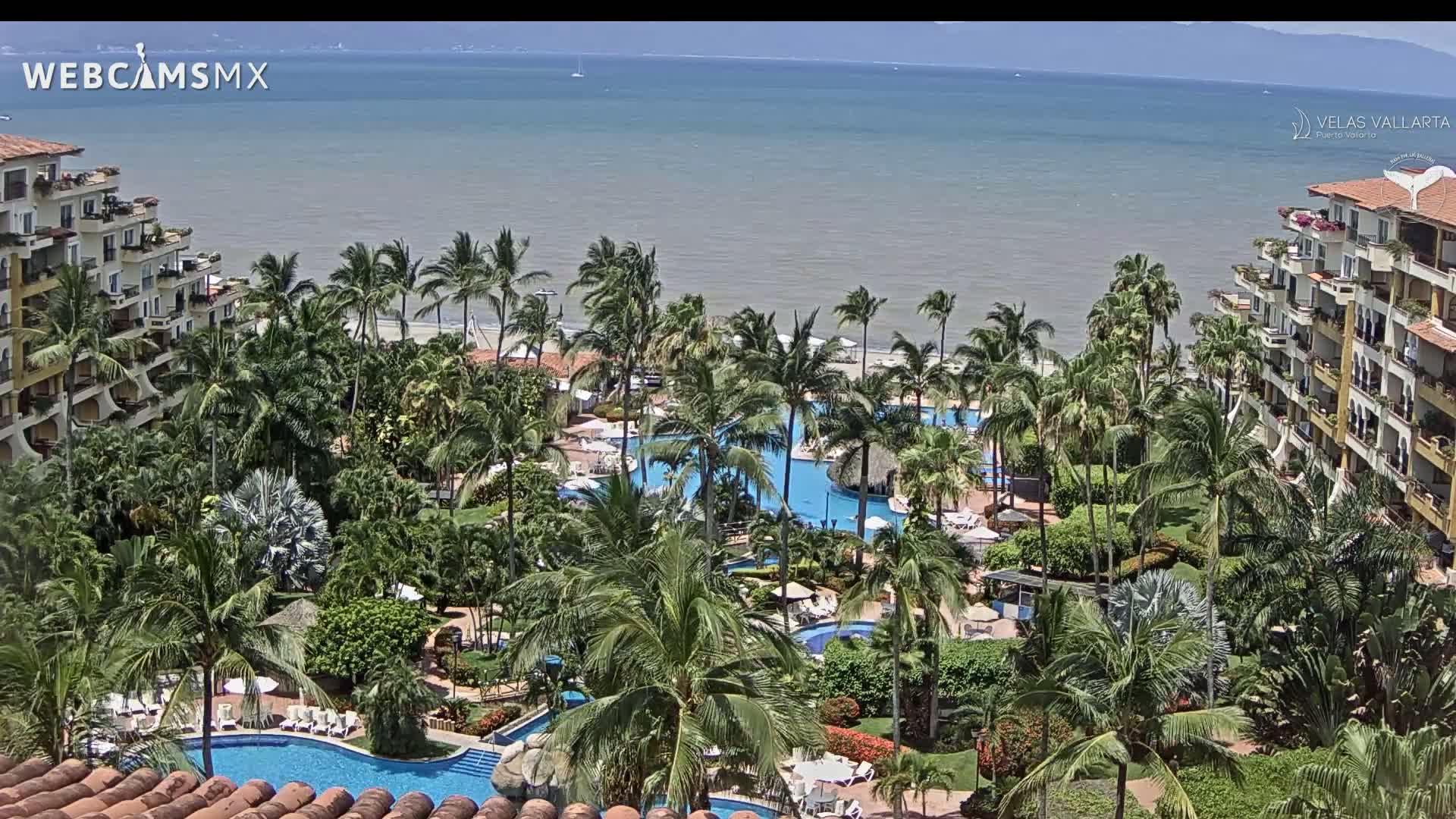 A resort with multiple pools surrounded by lush tropical landscaping overlooks a calm ocean under a sunny sky.