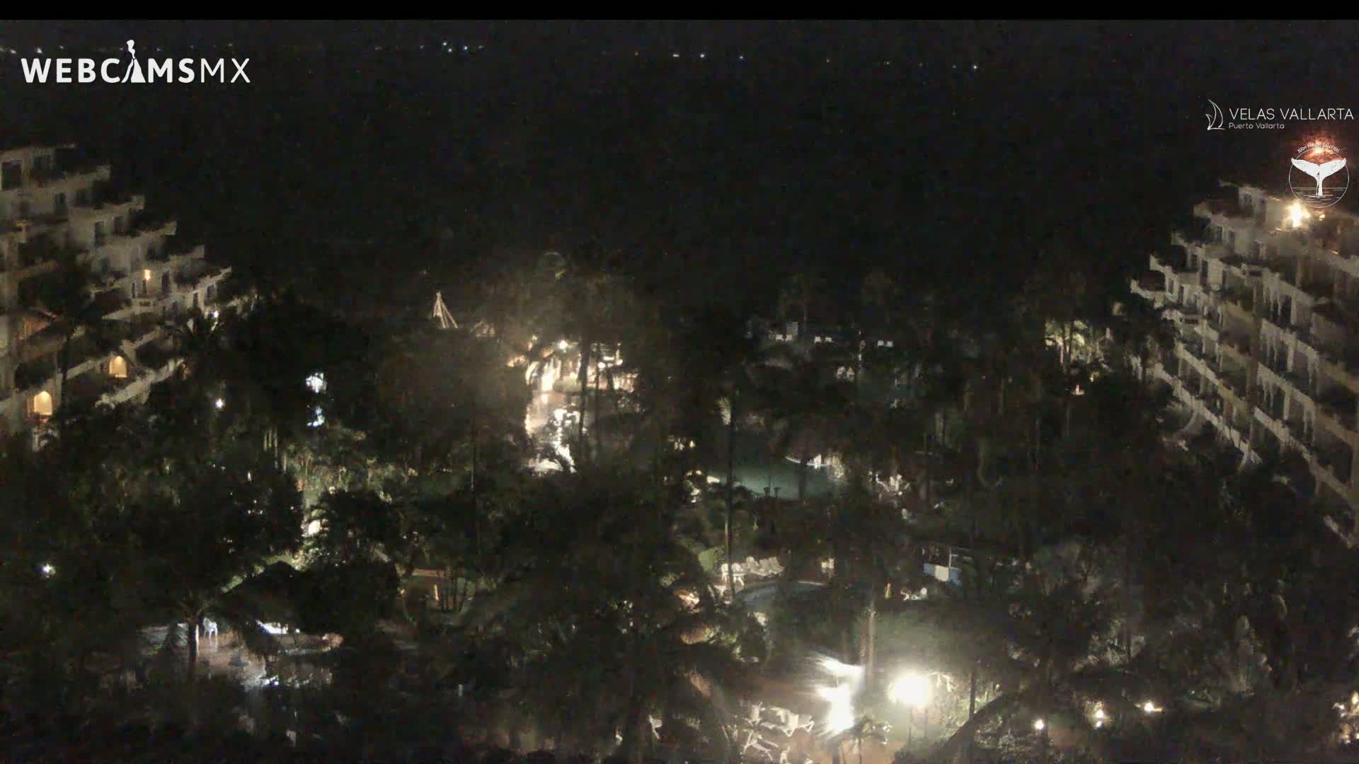 A nighttime, high-angle view of a resort complex with pools and palm trees, illuminated by scattered lights.