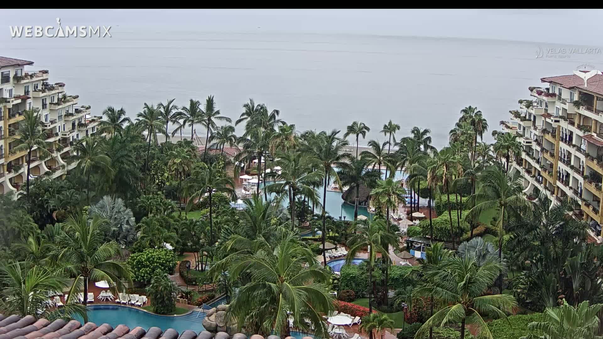 A resort with multiple pools surrounded by palm trees and buildings overlooks a calm ocean under an overcast sky.