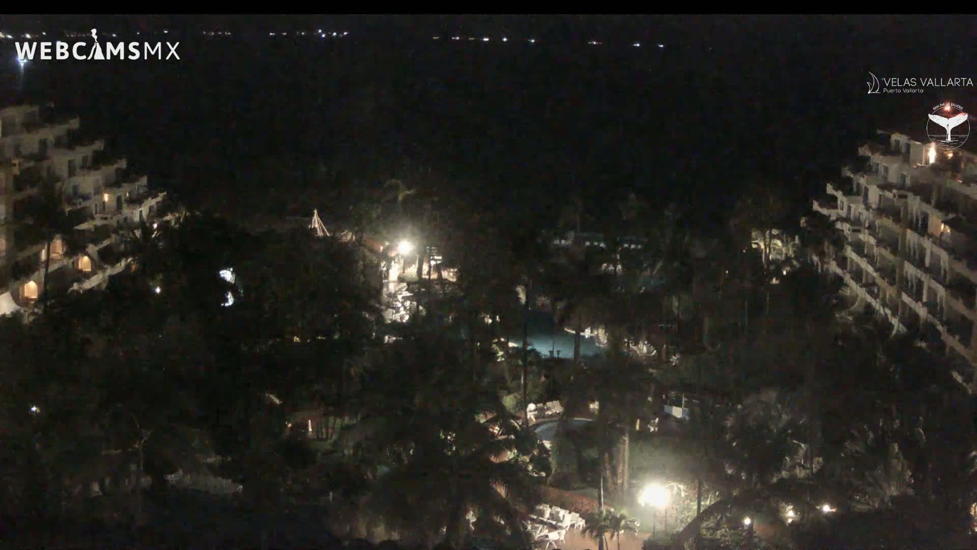A nighttime aerial view shows a resort complex with buildings, pools, and lush landscaping under a clear, dark sky.
