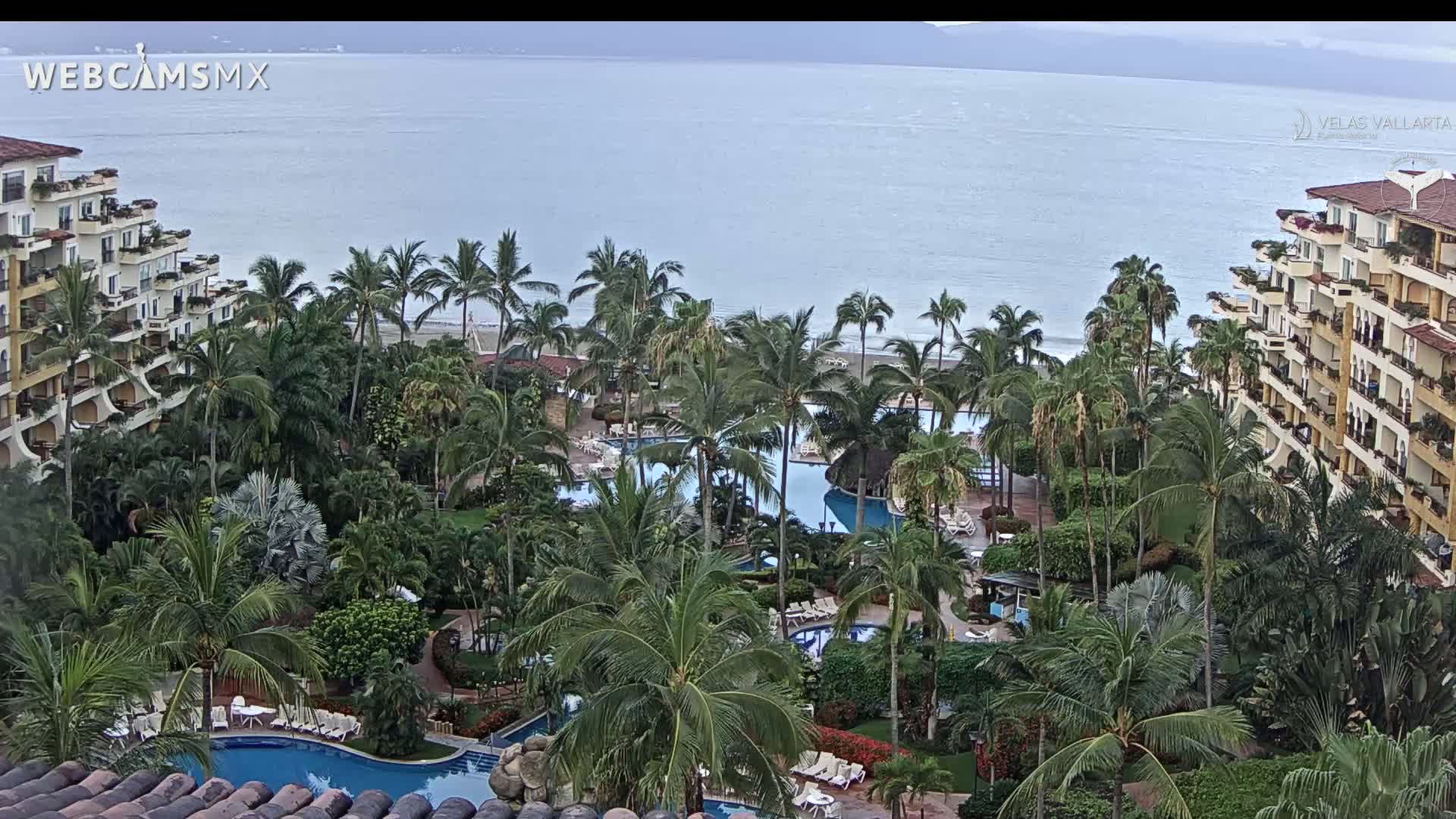 An overcast day reveals a tropical resort with multiple pools surrounded by lush palm trees and buildings overlooking a calm ocean.