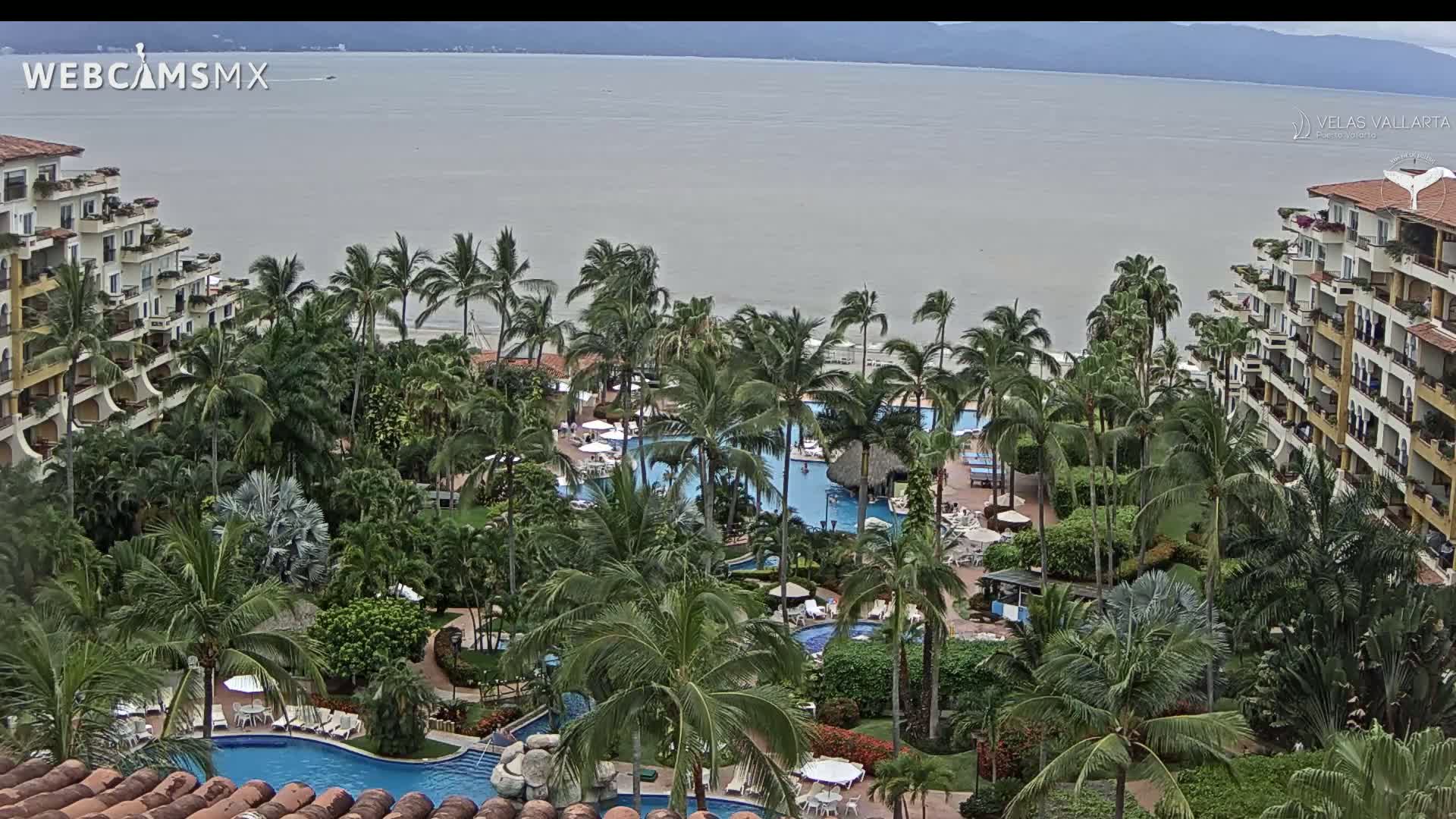 An aerial view of a tropical resort with multiple pools surrounded by palm trees and buildings, overlooking a calm ocean under an overcast sky.