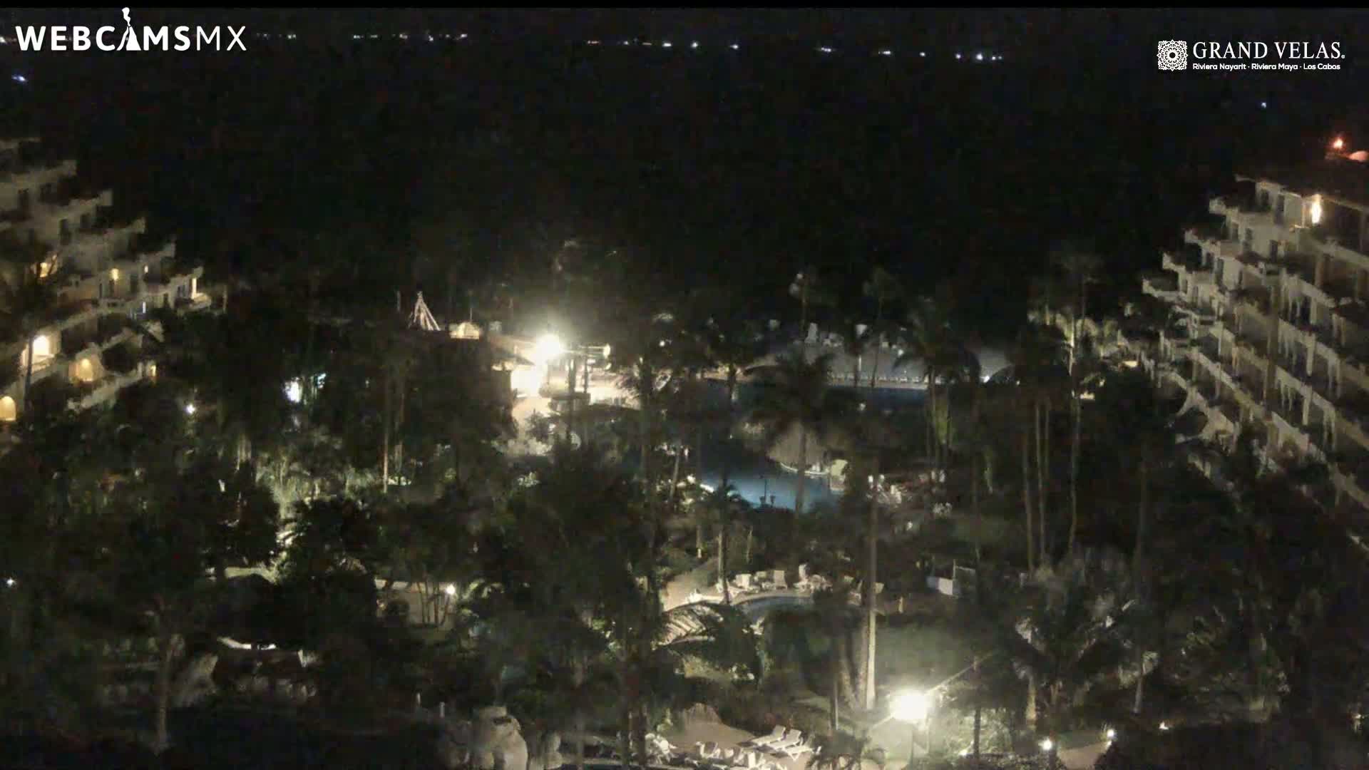 A nighttime view of a resort with multiple buildings, lush tropical landscaping, and illuminated pools.