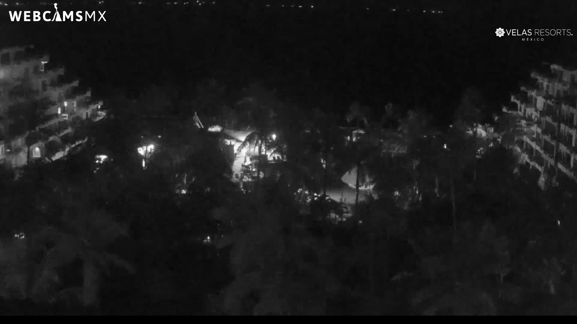 A black and white nighttime view shows an illuminated resort complex with buildings flanking a central area dense with palm trees and outdoor lighting, all under a clear night sky.