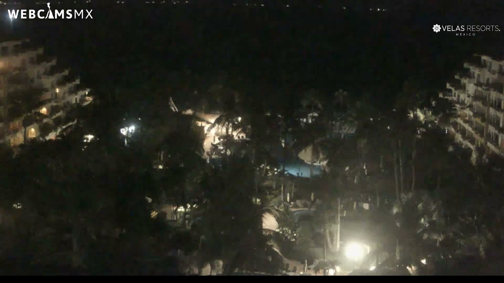 The image displays a sprawling resort complex at night under clear skies, illuminated by artificial lights revealing multi-story buildings, numerous palm trees, and a pool area.