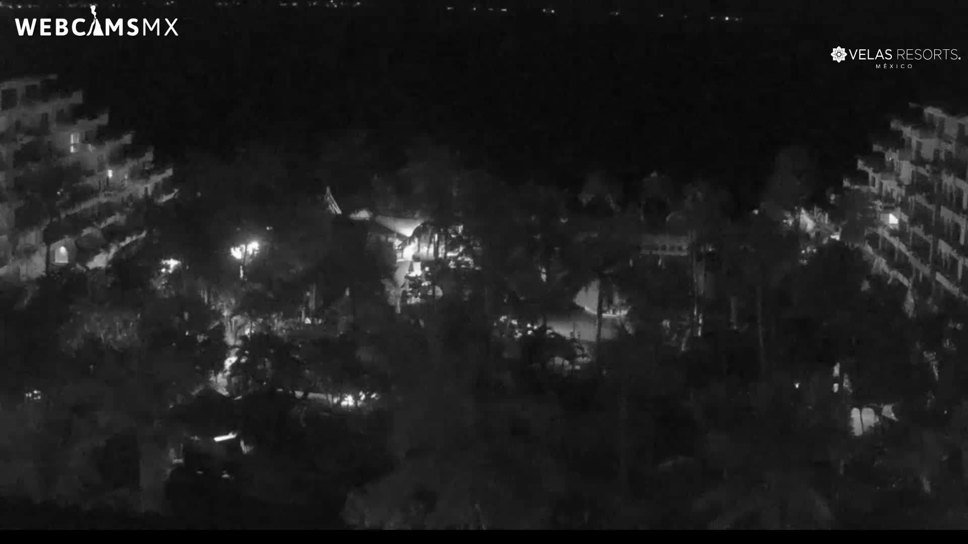 The image shows a clear night view of a resort complex with illuminated multi-story buildings on the left and right, framing a central area dense with trees and subtle lights.