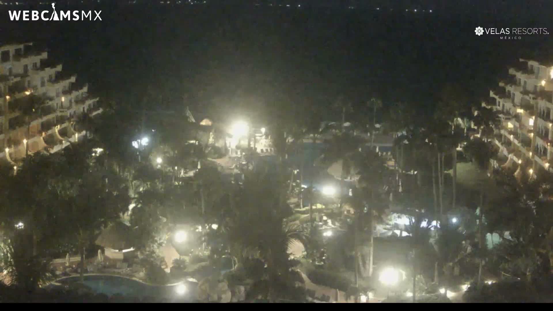 An elevated nighttime view reveals a brightly lit tropical resort complex featuring multiple buildings, palm trees, and swimming pools under a clear, dark sky.