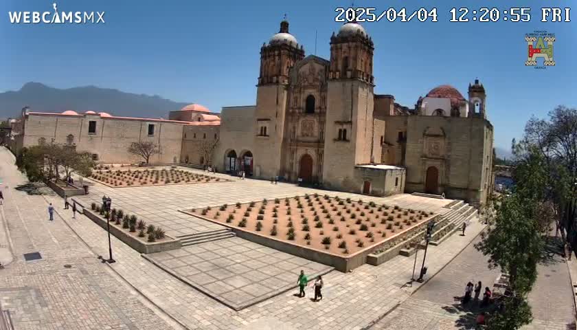 A large stone church with twin towers stands in a plaza with a geometrically planted garden, under a clear blue sky.