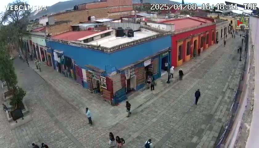 A cobblestone street scene under a mostly clear sky shows a row of brightly colored buildings, with several people walking along the street and near the buildings.