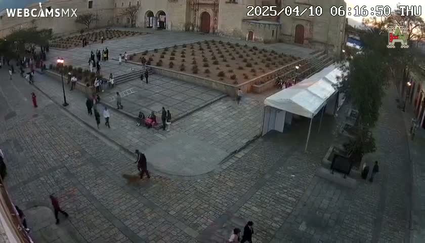 A high-angle, outdoor view shows a cobblestone plaza with a few people scattered throughout, a central garden, and a large white tent, under a partly cloudy sky.