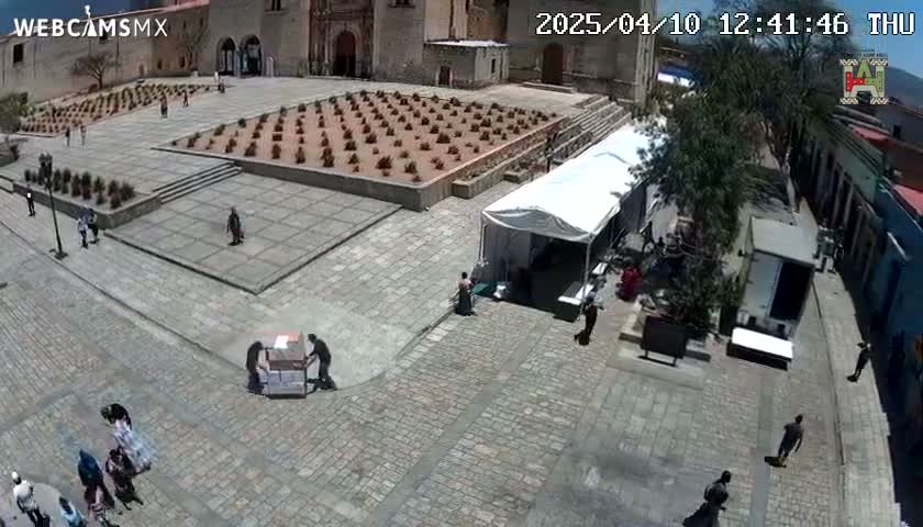 A high-angle view shows a cobblestone plaza on a sunny day, with people walking around, a few vendors setting up stalls, and a large building in the background.