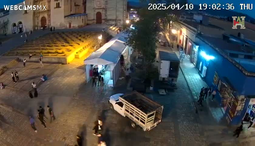 An aerial nighttime view of a town square with people walking around, a few vehicles parked, and a long white tent set up, under a mostly clear sky.