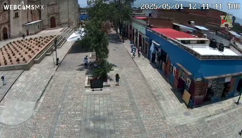 A cobblestone plaza and street scene, viewed from above on a sunny day, shows a few pedestrians near buildings and a planted garden.