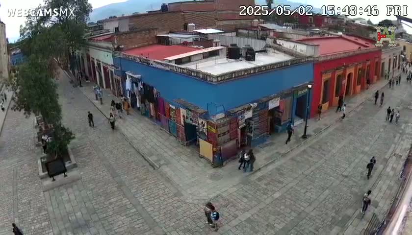 A cobblestone street scene shows pedestrians walking past colorful buildings under a clear sky.