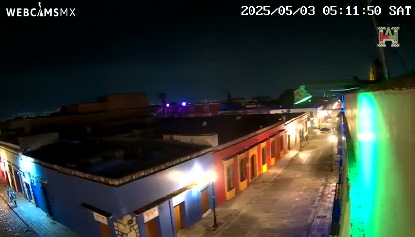 A nighttime, high-angle view shows a brightly lit street lined with colorful buildings under a clear, dark sky.