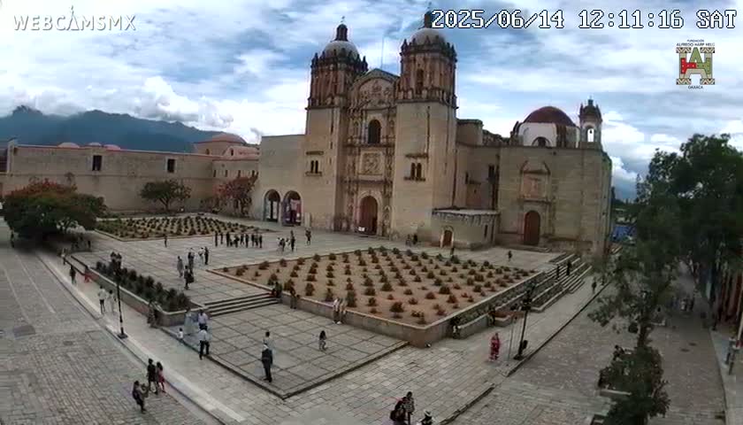 A large church complex with a courtyard and surrounding buildings is shown on a partly cloudy day with mountains in the background.