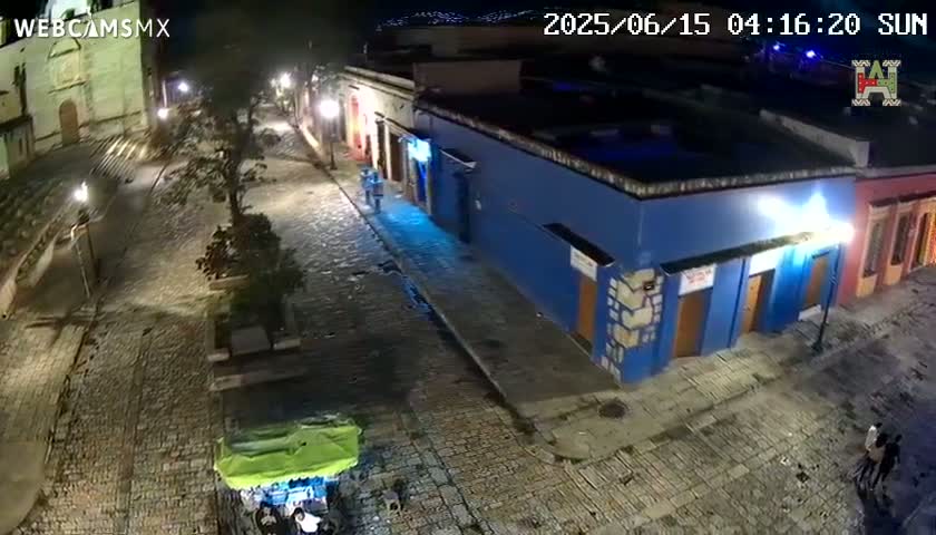 A nighttime, high-angle view shows a cobblestone street lined with buildings, one of which is painted blue, with a few people walking and a small vendor stand present.