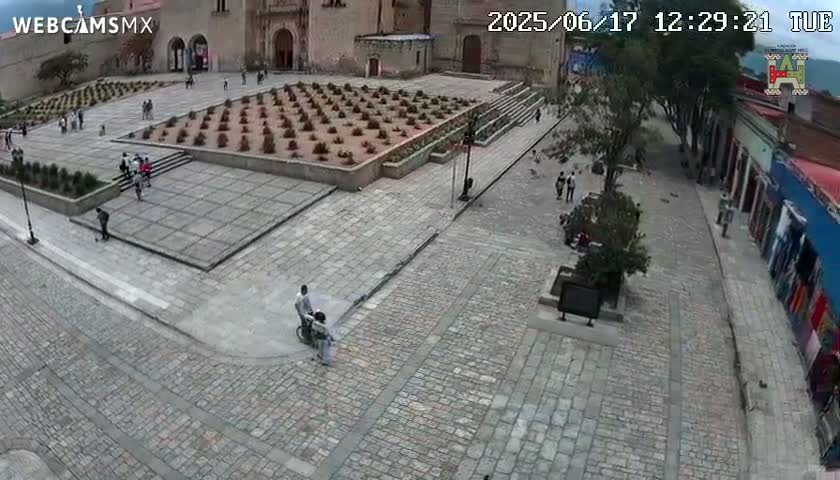 A cobblestone plaza with a central garden, a stone building, and sparsely populated streets is seen from above on a clear day.