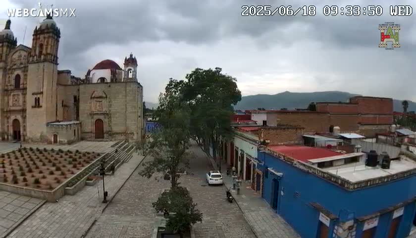 A large stone church stands in a town square under an overcast sky, with a cobblestone street leading to other buildings, including a blue one.