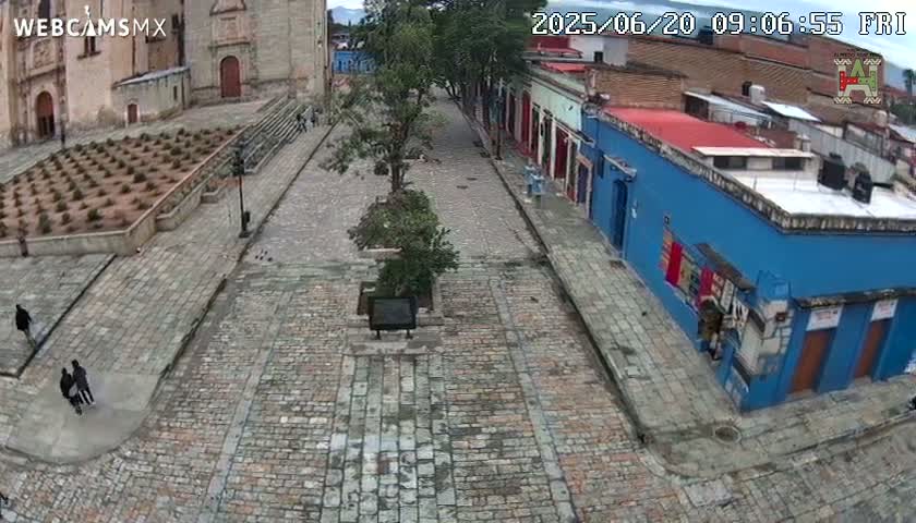 A cobblestone plaza, bordered by colonial-style buildings, with a few pedestrians and small trees, under an overcast sky.