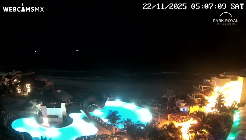 An aerial night view reveals a brightly lit resort with glowing blue swimming pools and illuminated buildings situated along a dark ocean shore, under a clear night sky.