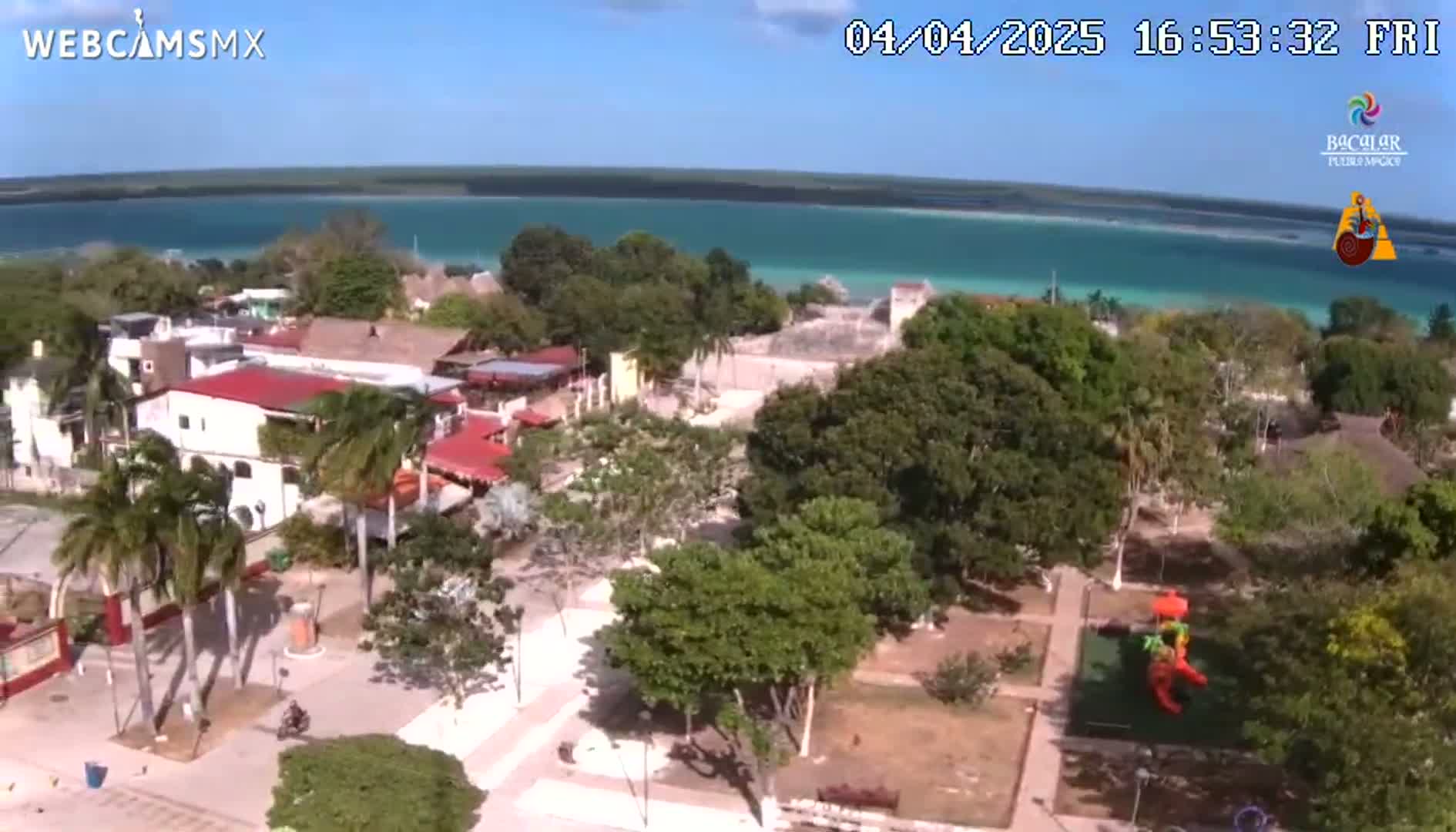 An elevated view shows a town's plaza and buildings next to a bright turquoise body of water under a clear blue sky.