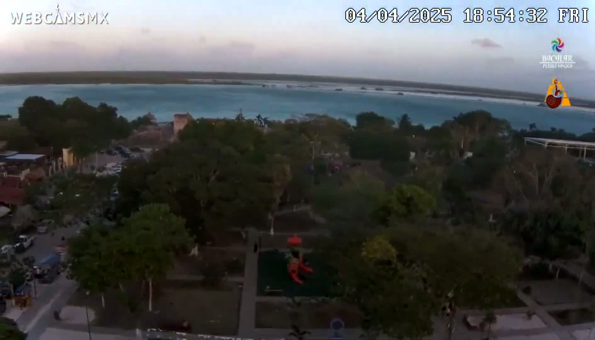 An aerial view shows a town with lush green trees and a playground near a calm, turquoise body of water under a partly cloudy, twilight sky.