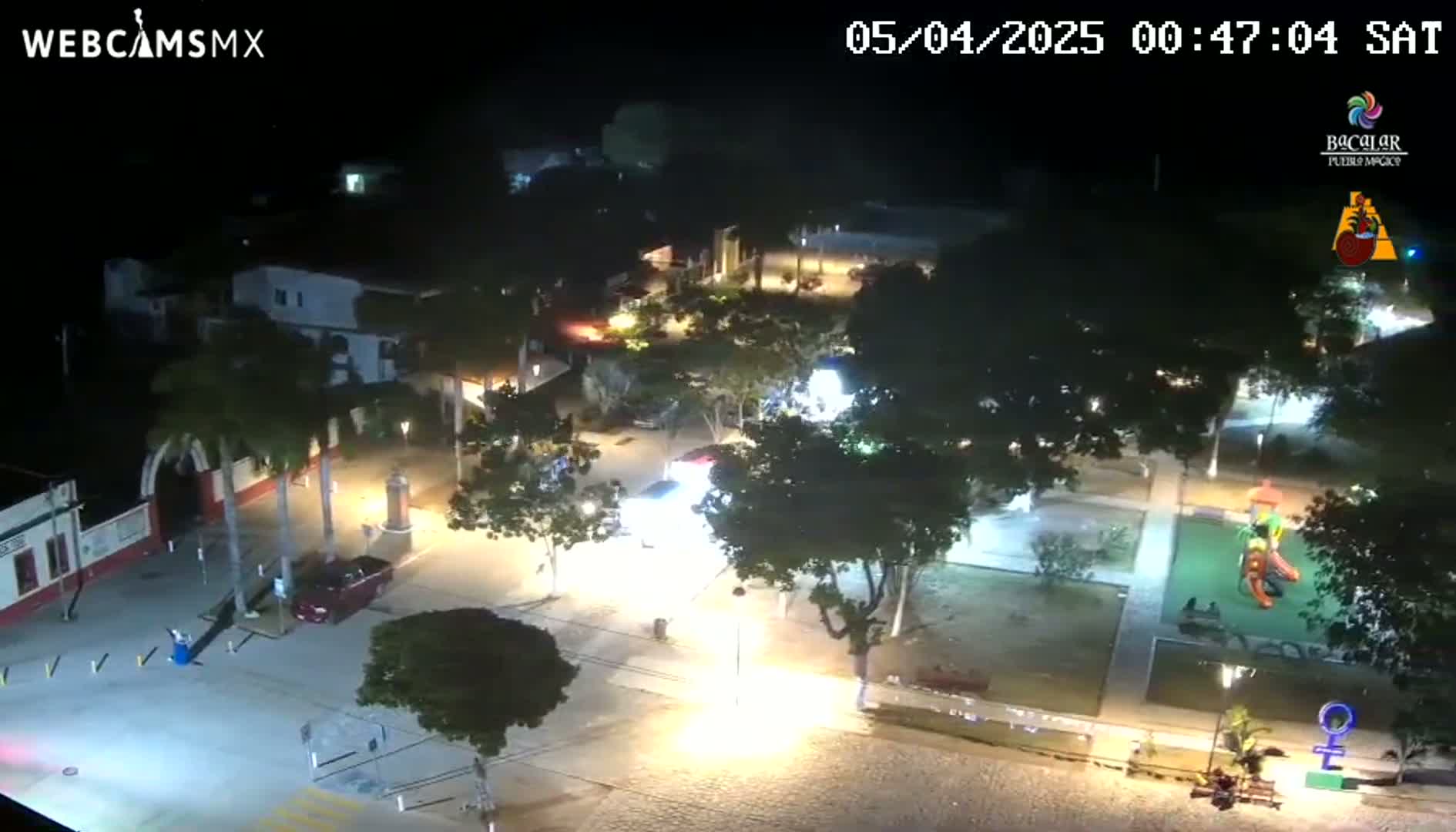 A nighttime aerial view of a town square with cars parked, trees, and a playground, under clear skies.