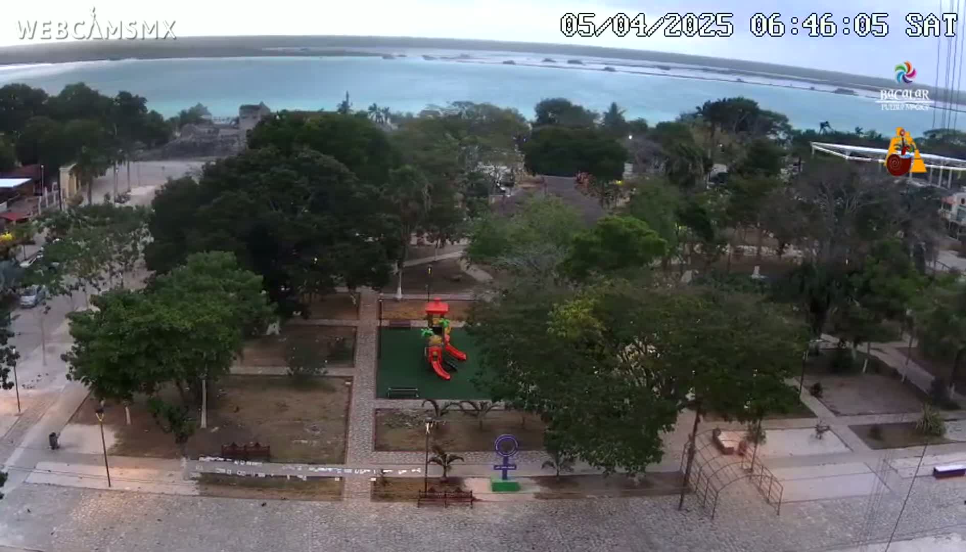 An aerial view shows a park with a playground, surrounded by trees and buildings, overlooking a calm body of water under a mostly clear sky.