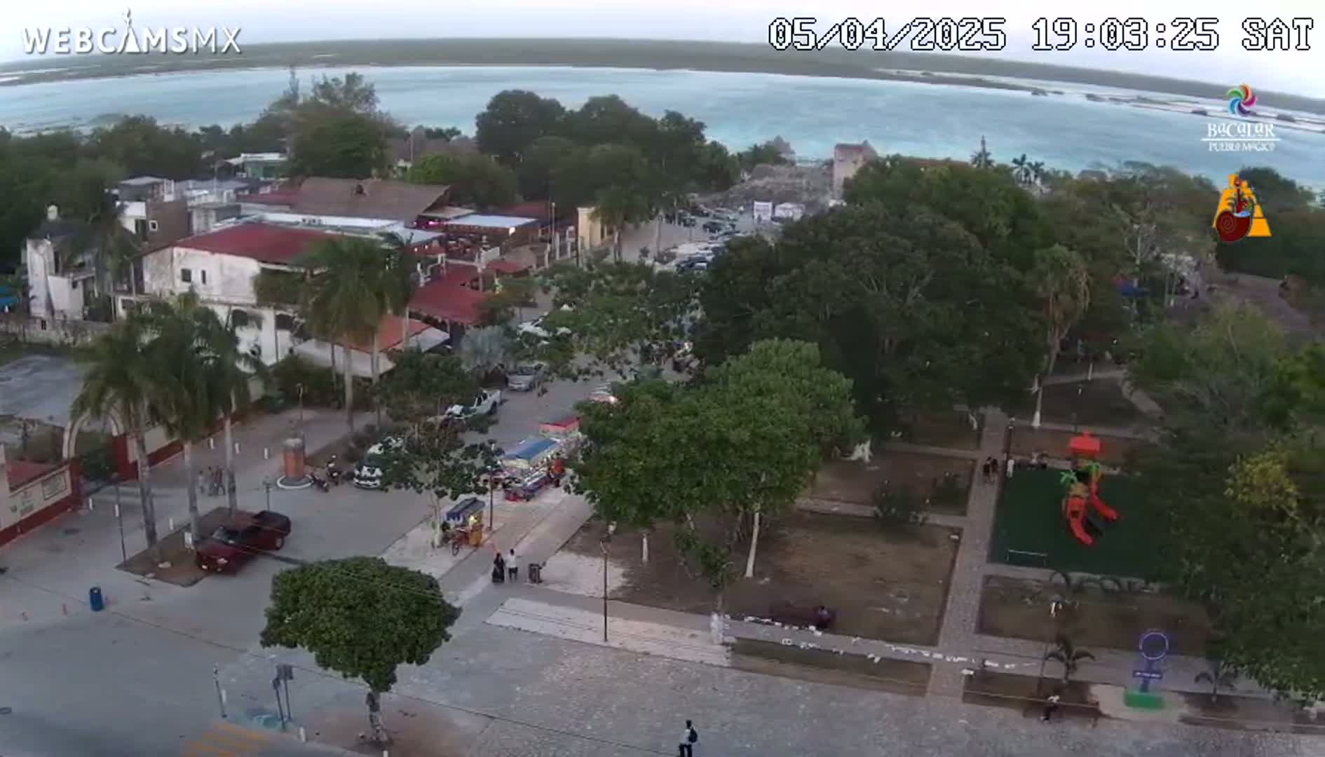 An aerial view of a town square near a body of water at dusk, with palm trees, buildings, and a playground visible.
