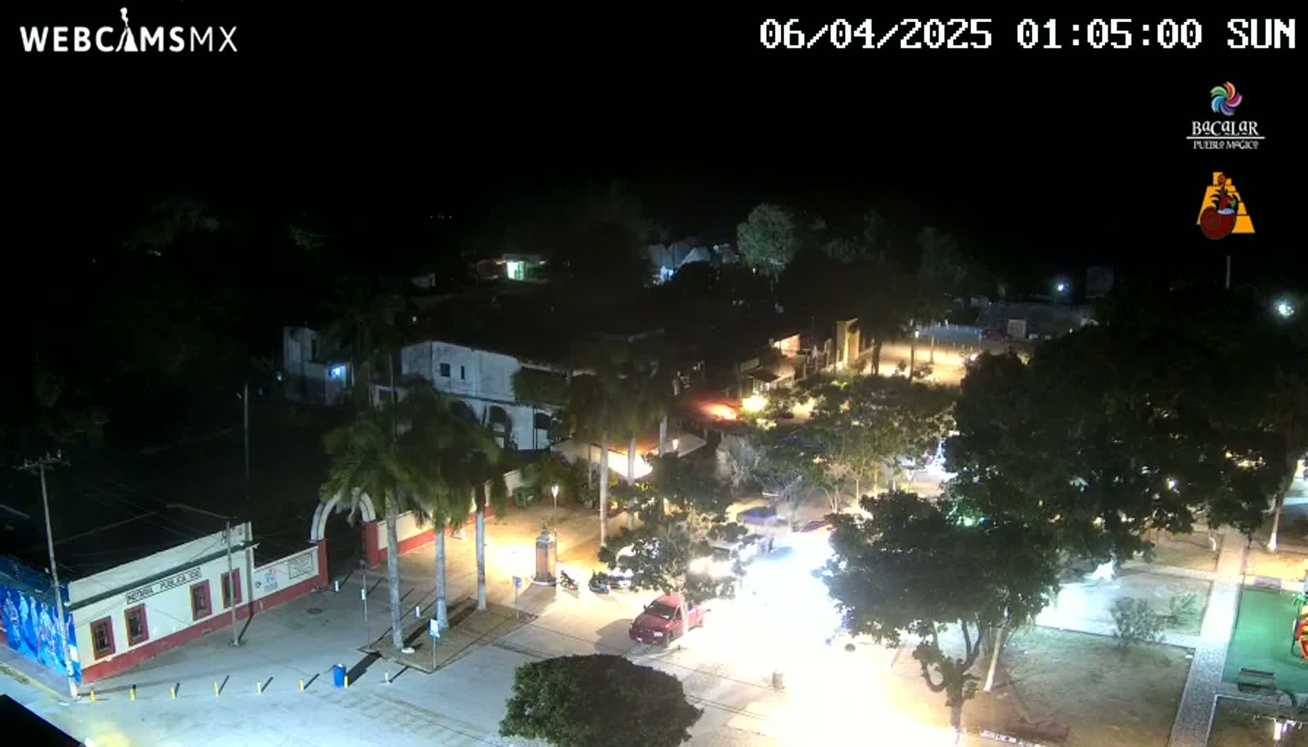 An aerial nighttime view of a town square with parked cars, illuminated buildings, and palm trees under clear skies.